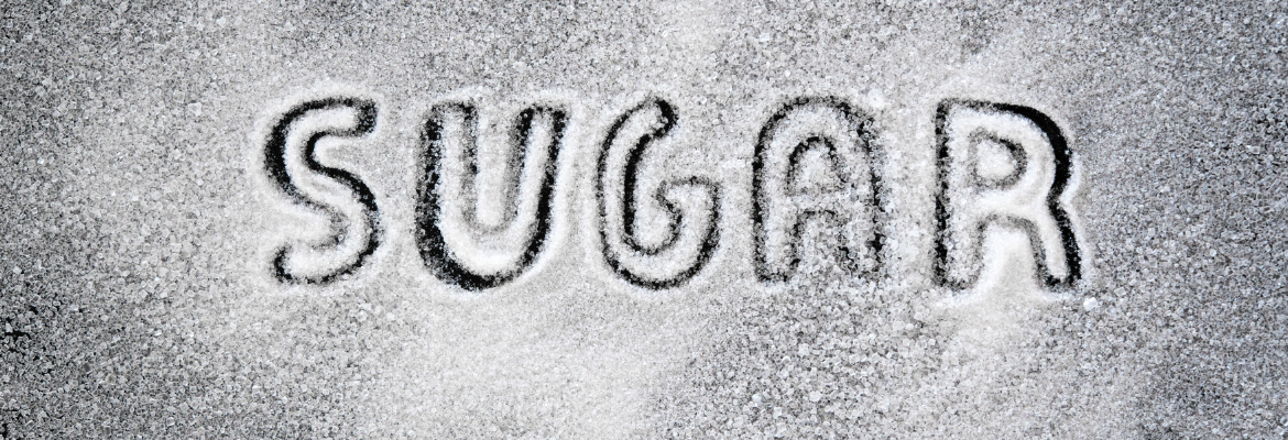 Picture shows sugar particles spread on a table and "Sugar" inscribed on it with fingers.